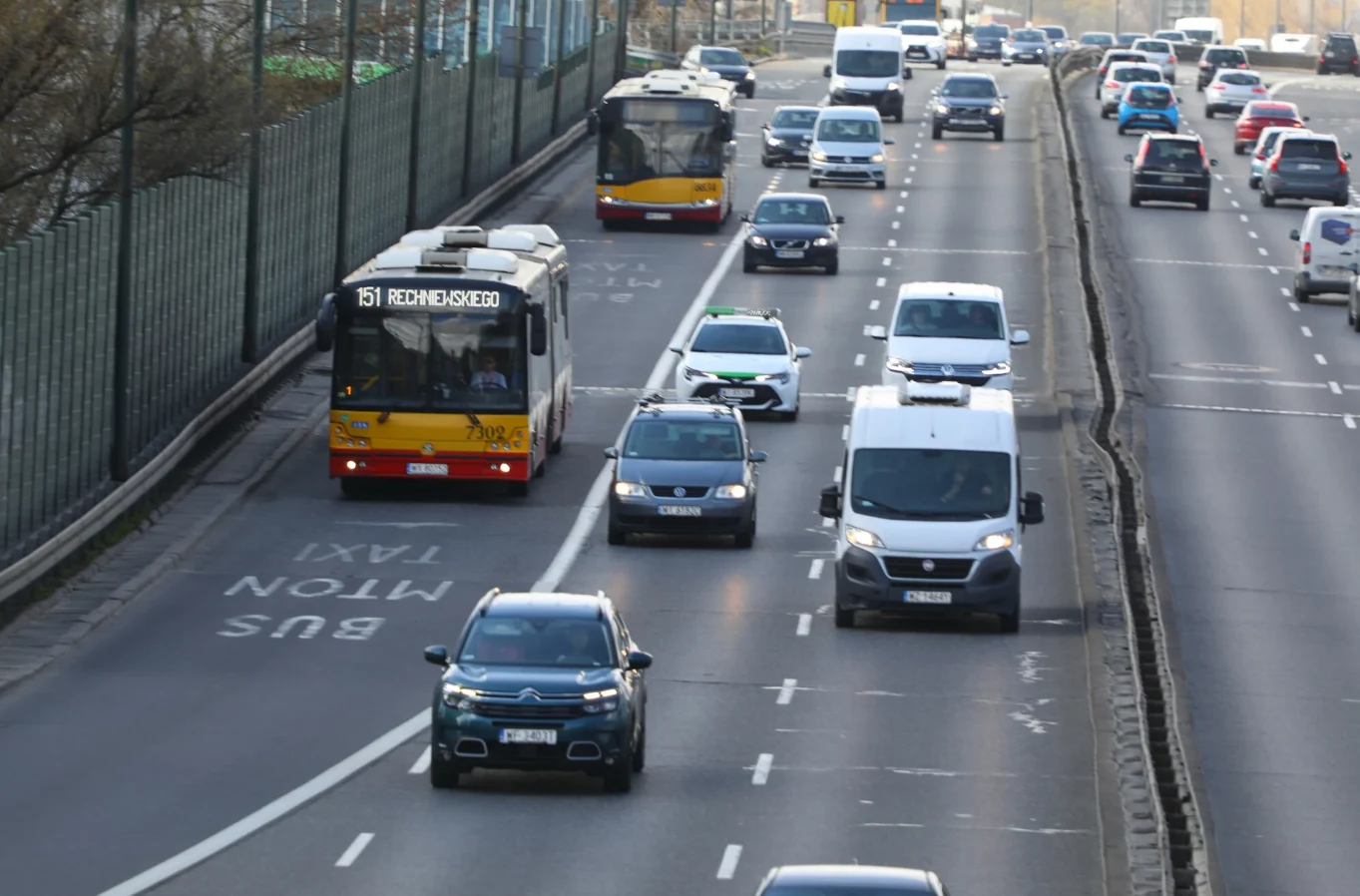 Zatłoczona autostrada z buspasem, na którym jedzie autobus. Pozostałe pasy zajęte przez auta osobowe i dostawcze. W tle infrastruktura drogowa i bariera dźwiękochłonna.