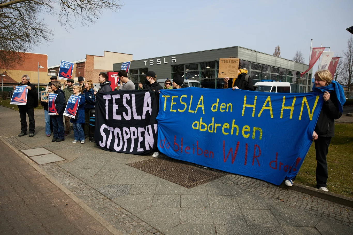Grupa ludzi stoi przed budynkiem z logo Tesla, trzymając bannery z napisami "TESLA STOPPEN" i "TESLA den HAHN abdrehen, bleiben WIR dran". W tle widać zaparkowane samochody i budynek z napisem Tesla.