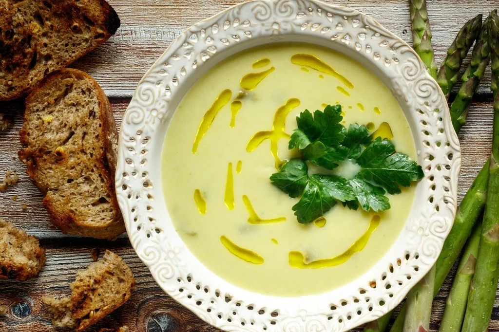 Zupa szparagowa Kremowa zupa ze świeżych szparagów z dodatkiem oliwy i pietruszki, podana w ozdobnym talerzu. Obok leżą kromki chrupiącego chleba oraz świeże szparagi na drewnianym stole.