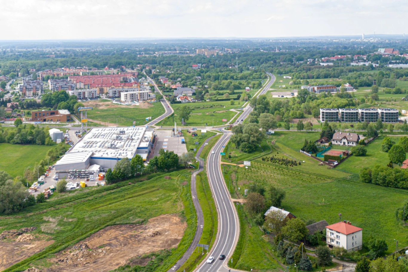 Budowa obwodnicy Oświęcimia wkracza w kluczowy etap. Widok z lotu ptaka na rozległy teren podmiejski z zielonymi łąkami i polami. Widoczna główna droga biegnie przez środek, otoczona budynkami mieszkalnymi i przemysłowymi. Liczne domy są rozproszone na tle zieleni.
