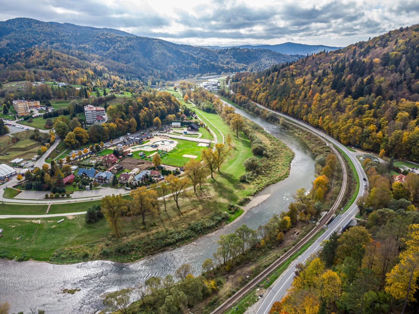 Malowniczo położona Muszyna to uzdrowisko z bogatą ofertą przez cały rok. Krajobraz górski z wijącą się rzeką, otoczony kolorowymi jesiennymi drzewami. Widoczne są budynki i drogi biegnące wzdłuż doliny, podkreślające harmonijną współpracę natury i architektury.