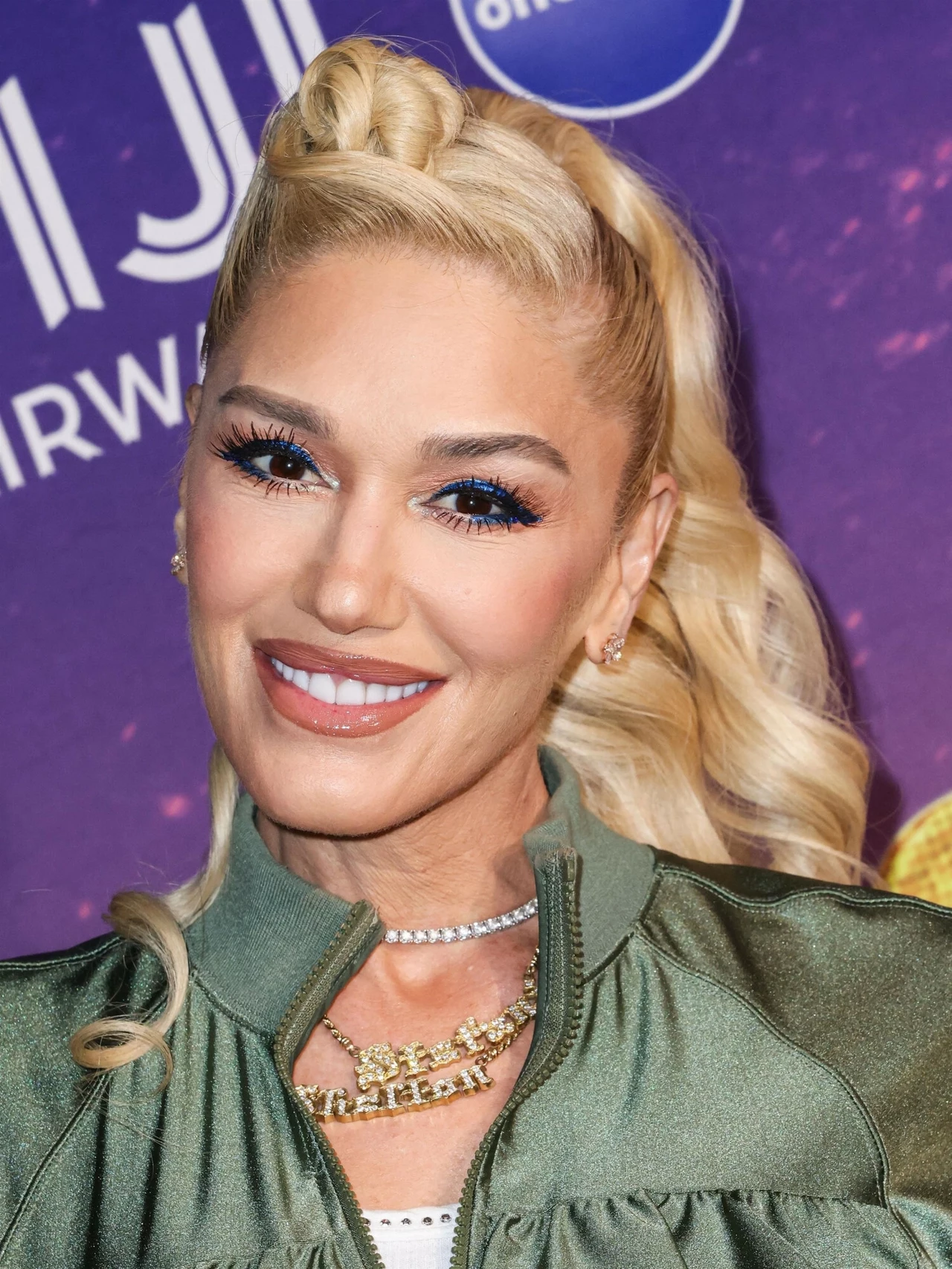 Gwen Stefani zachwyca świetną formą i młodym wyglądem Uśmiechnięta kobieta z blond włosami ułożonymi w efektowną fryzurę, w zielonej kurtce i naszyjniku. Na tle fioletowej ściany z elementami graficznymi.