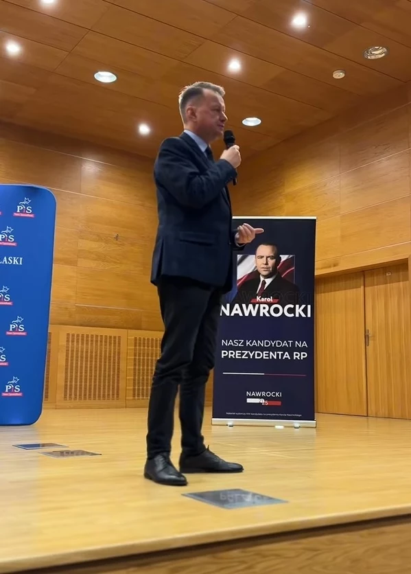 Mariusz Błaszczak na spotkaniu w Białej Podlaskiej. W tle plakat Karola Nawrockiego Mężczyzna w garniturze przemawia do publiczności z podium, trzymając mikrofon. W tle znajduje się plakat kandydata na prezydenta, na którym widnieje jego zdjęcie oraz nazwisko 'Nawrocki'. Na scenie i plakacie widnieje logo partii PIS.