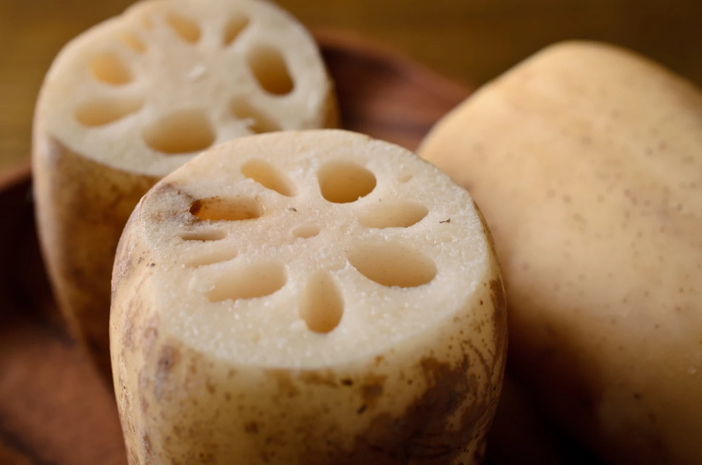 Korzeń lotosu pasuje zarówno do dań wegetariańskich, jak i mięsnych Korzeń lotosu pokrojony na plastry, z widocznym charakterystycznym wzorem otworów. Początki rośliny mają jasnobrązowy kolor z odrobiną ziemi. Poszczególne kawałki są ułożone na drewnianej tacy.