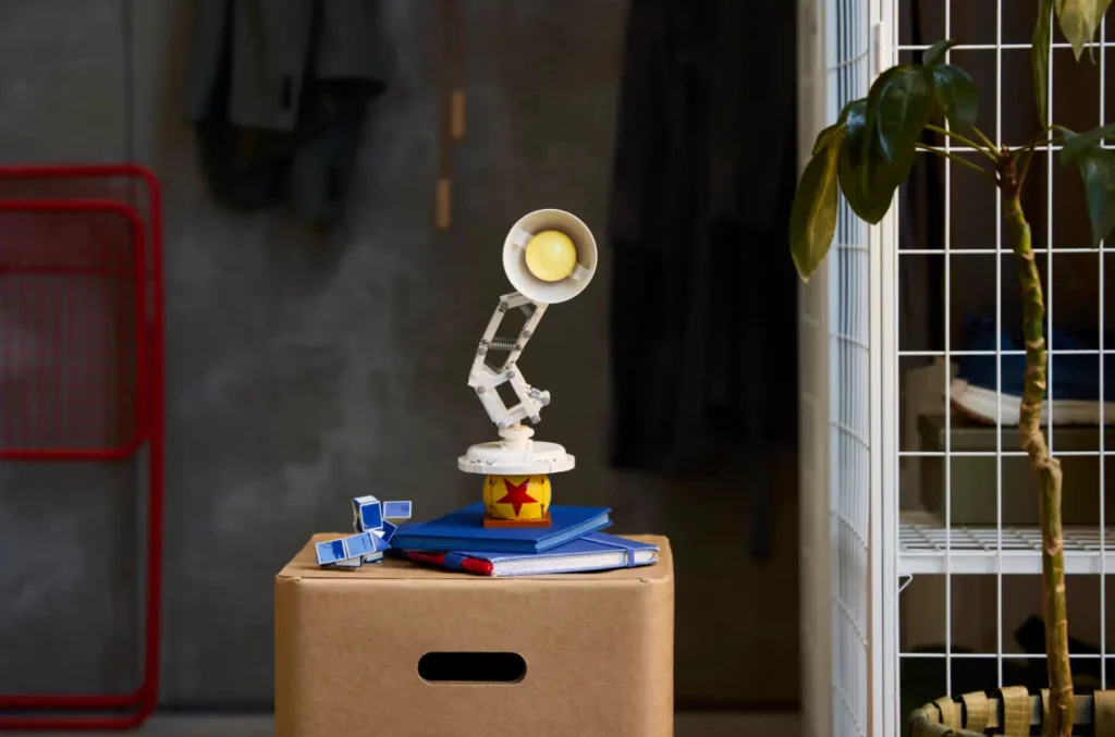 Lampka Luxo Jr. na piłce z gwiazdą – scena z filmu odtworzona z LEGO Na tekturowym pudełku stoi stylizowana lampka biurkowa z żarówką, przypominająca postać z animacji. Obok leżą niebieskie książki i niebieski kubik. W tle ściana z ubraniami i roślina w rogu.