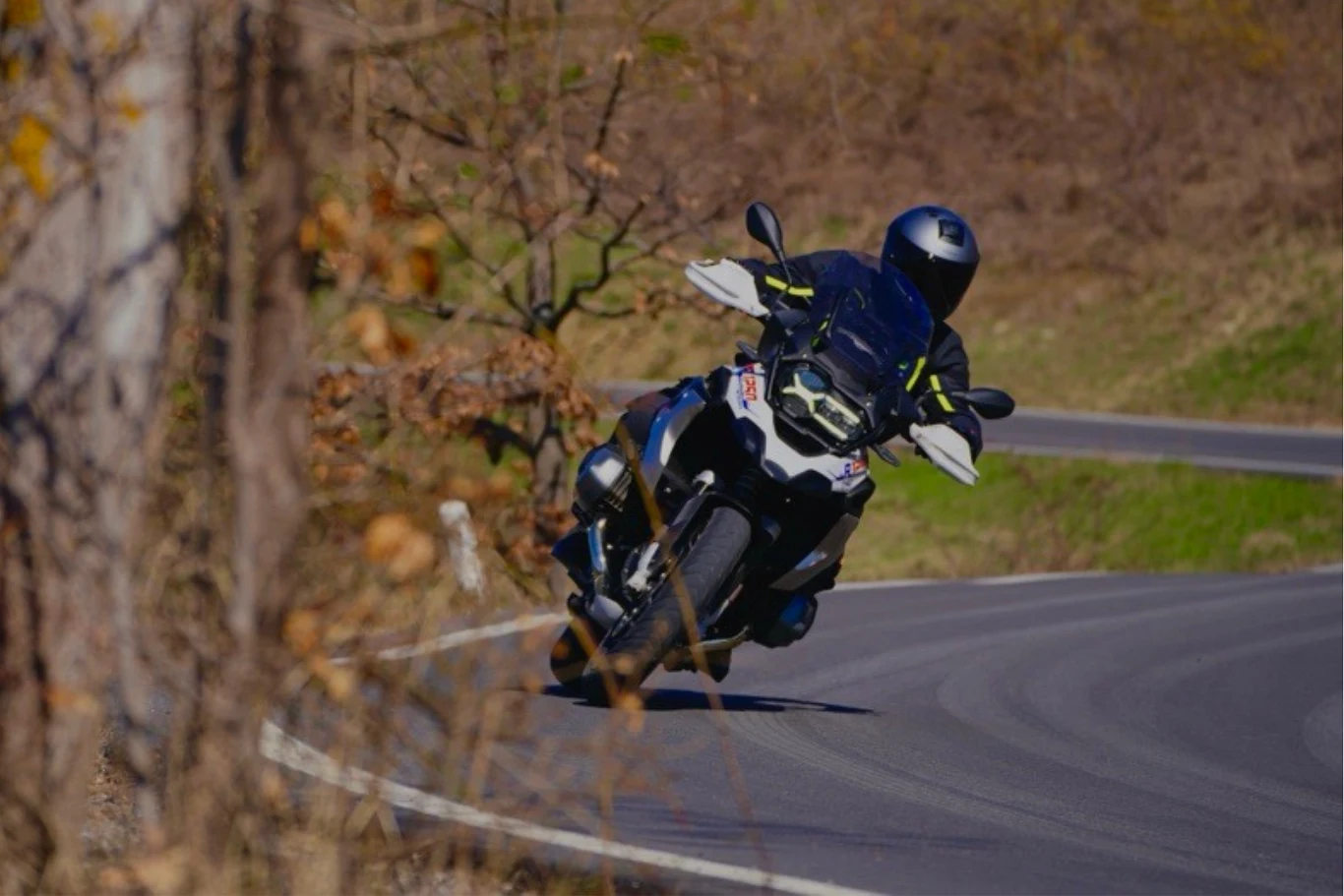 Wytrącenie motocykla z punktu równowagi jest kluczowe, aby pochylić go w stronę zakrętu. Niezbędne do tego jest skorzystanie z przeciwskrętu. Motocyklista w kasku pokonuje zakręt na krętej, asfaltowej drodze. W tle widoczna jest jesienna sceneria z drzewami o brązowych i żółtych liściach. Motocykl jest dynamicznie przechylony na zakręcie, co podkreśla szybkość jazdy.