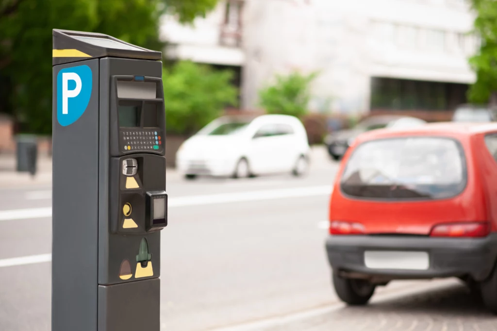 Mecenas nie zdążyła przedłużyć postoju na płatnym parkingu. Automat parkingowy na chodniku, obok zaparkowane czerwone auto. W tle inne pojazdy i rozmazana zieleń.