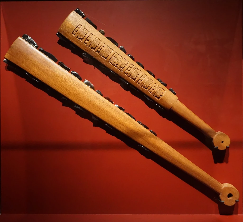 Rekonstrukcja macuahuitl Dwie drewniane pałki ceremonialne na tle czerwonej ściany, ozdobione wzorami i symbolami co wskazuje na ich kulturowe znaczenie.