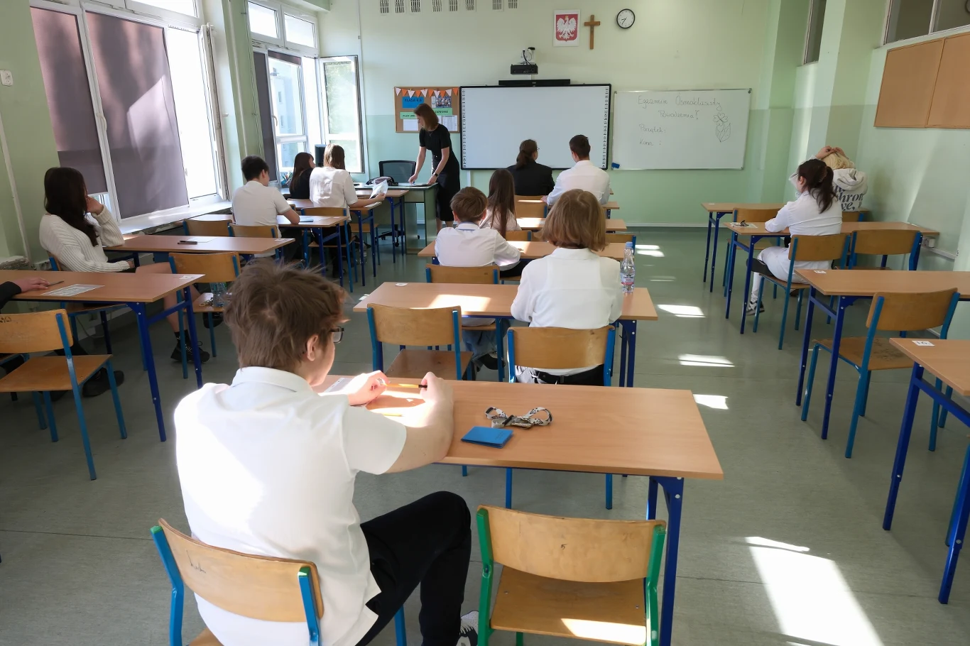 Język polski na egzaminie ósmoklasisty. Polonistka komentuje Uczniowie w szkolnej klasie siedzą w rzędach przy biurkach. Na przodzie znajduje się nauczyciel przed tablicą, prowadzący lekcję. Ściany ozdobione są godłem Polski oraz krzyżem, a na tablicy widoczne są notatki z lekcji.