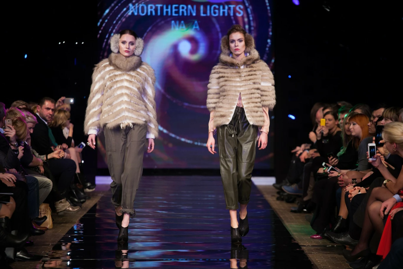Dwie modelki prezentujące luksusowe futra idące po wybiegu na pokazie mody. W tle widoczny efekt świetlny przypominający zorzę polarną oraz napis 'Northern Lights'. Publiczność siedząca po obu stronach obserwuje pokaz.
