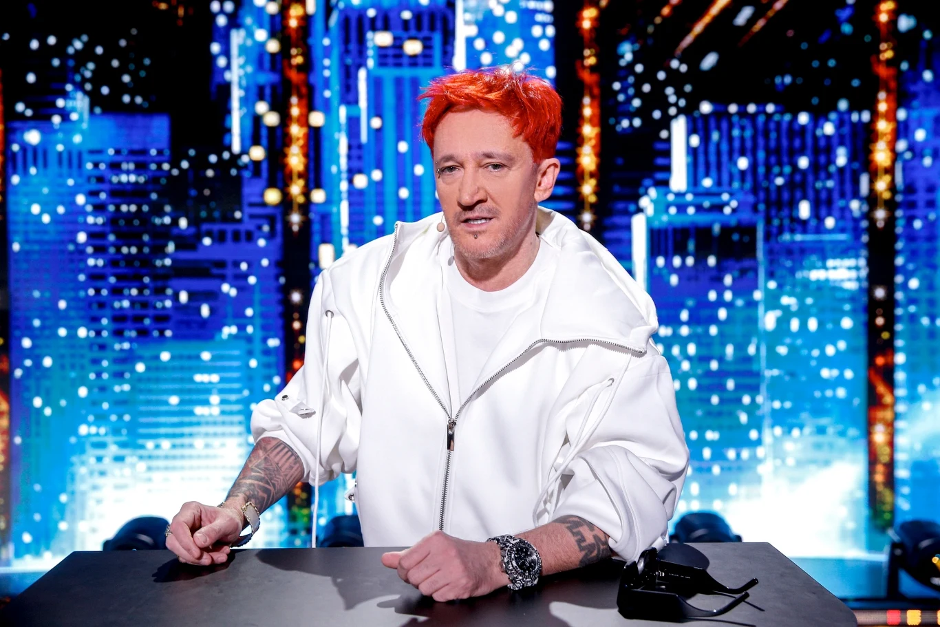 Michał Wiśniewski z grupą Ich Troje znalazł się w finale plebiscytu Złotego Bączka Mężczyzna w białej bluzie z pomarańczowymi włosami siedzi przy stole na tle świetlnych miastowych wieżowców.