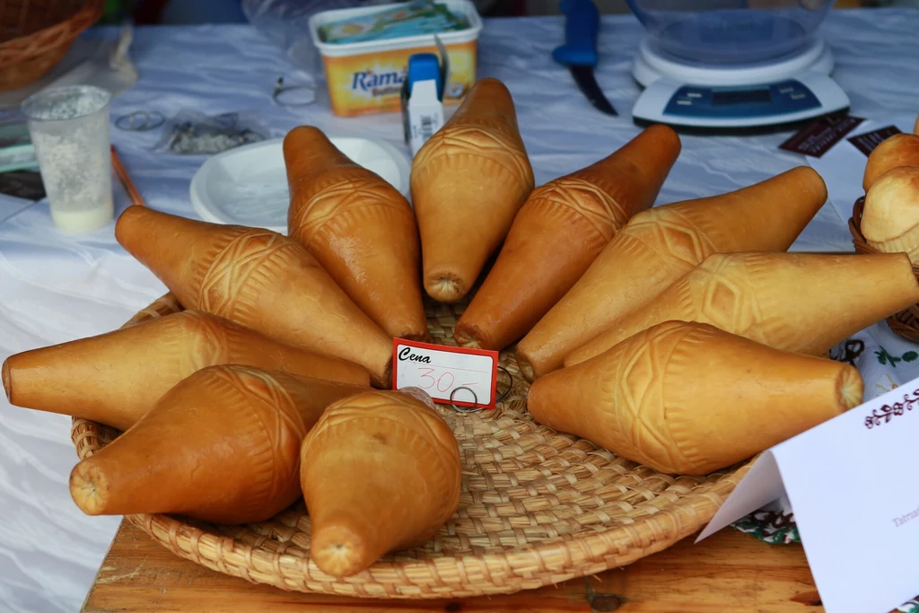 Tradycyjne, prawdziwe oscypki spełniające wymogi unijne Oscypki ułożone na wiklinowym koszu, obok cena na kartce papieru. W tle widoczne produkty spożywcze, takie jak margaryna, i naczynia na stole.