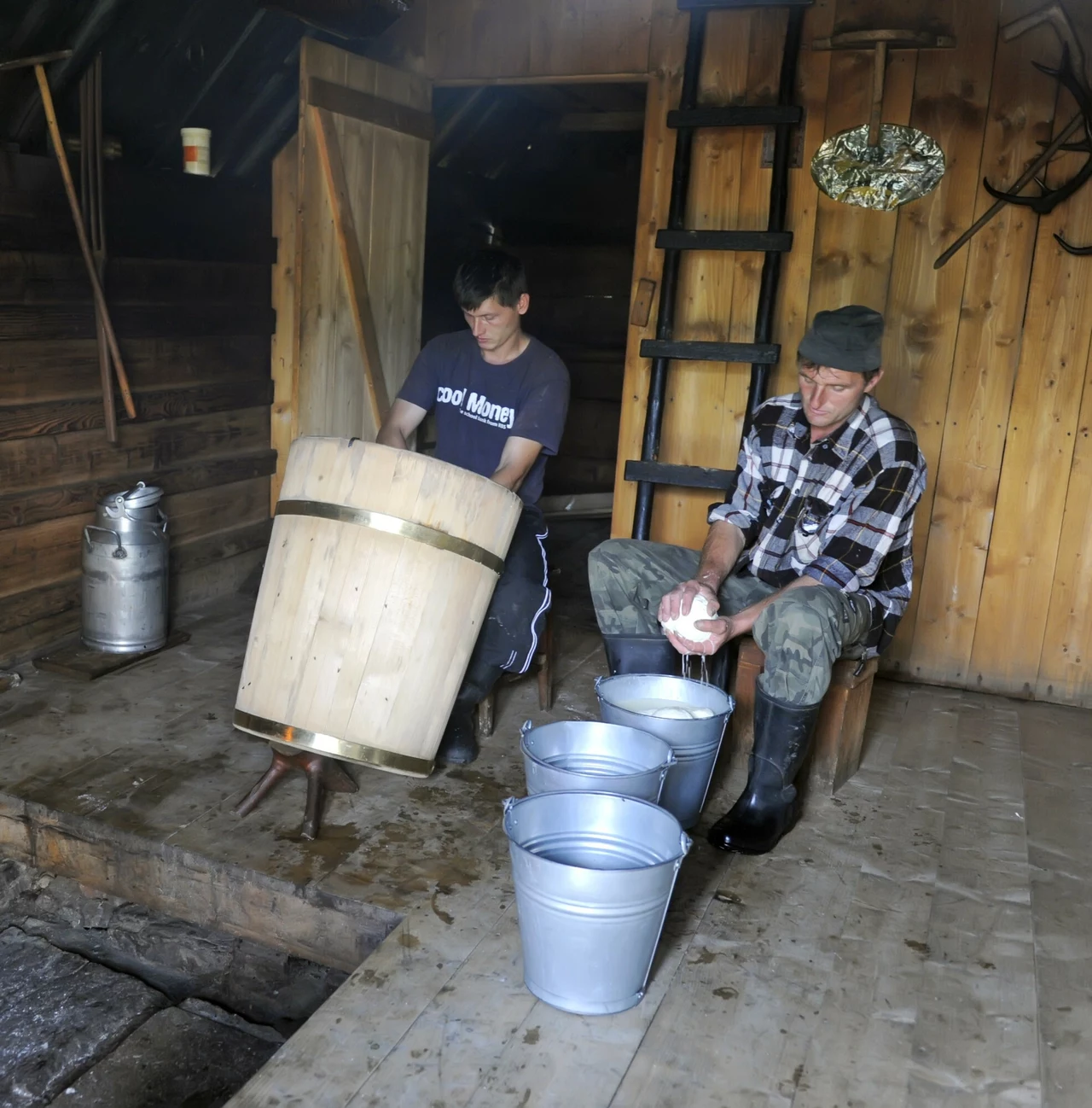 Górale wyrabiają oscypki w bacówce Dwóch mężczyzn w drewnianej chacie, jeden z nich siedzi na ławce i przygotowuje coś w drewnianym naczyniu, drugi siedzi, obiera ziemniaki lub inne warzywo do metalowych wiader ustawionych przed nim. W tle drabiną prowadząca na wyższy poziom.
