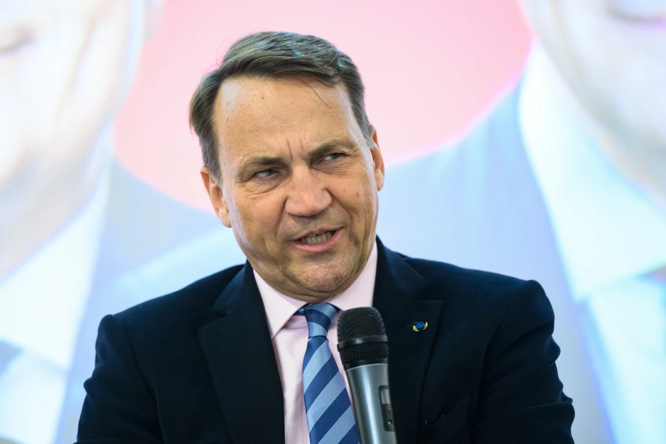 Minister spraw zagranicznych Radosław Sikorski podczas spotkania w Poznaniu Mężczyzna w garniturze z różowym krawatem, trzyma mikrofon i mówi. W tle rozmyty obraz innej postaci.