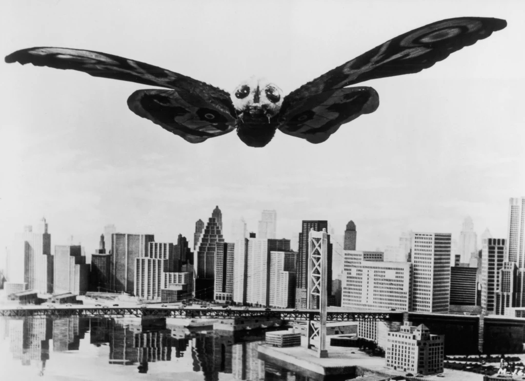 Japoński potwór Mothra, towarzysz Godzilli Ogromny, fantastyczny motyl unosi się nad miastem, dominując na tle nowoczesnych budynków i mostu. Scena przypomina widowiskowe efekty specjalne z filmów science fiction.