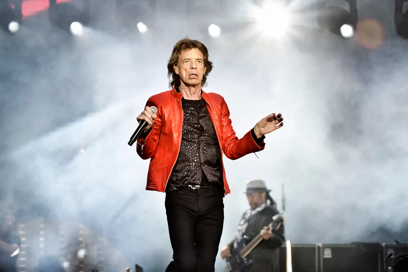 Mick Jagger z the Rolling Stones Lider zespołu rockowego na scenie w czerwonej kurtce z mikrofonem w ręku. Za nim widać gitarzystę i efekty świetlne podkreślające dynamiczną atmosferę koncertu.