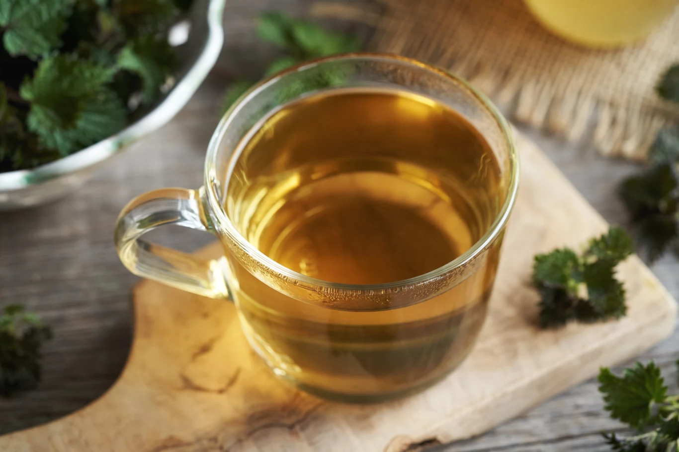 Pokrzywa świetnie nadaje się do przygotowania bogatego w witaminy naparu Szklanka pełna herbaty ziołowej na drewnianej desce, otoczona świeżymi liśćmi pokrzywy, co sugeruje naturalny i zdrowy napój.