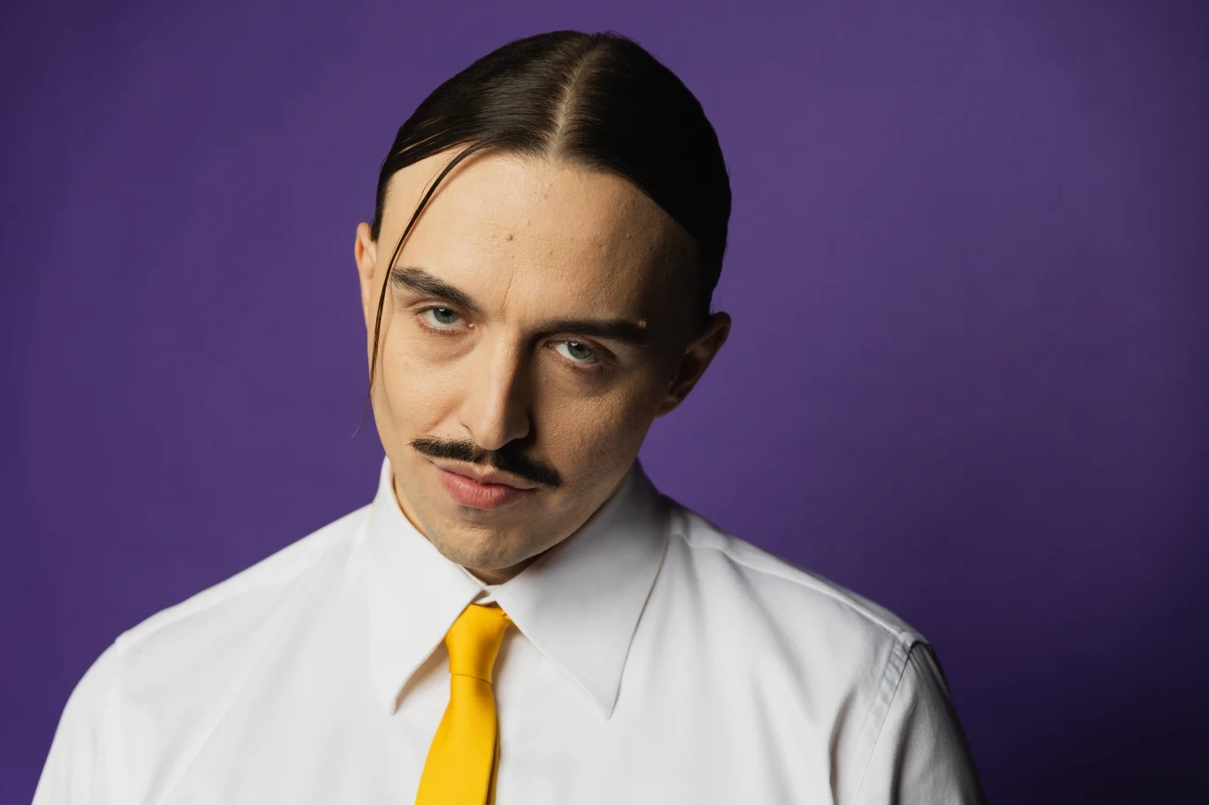 Tommy Cash - reprezentant Estonii w Konkursie Eurowizji 2025 Mężczyzna z wąsem i upiętymi włosami w białej koszuli i żółtym krawacie na fioletowym tle.