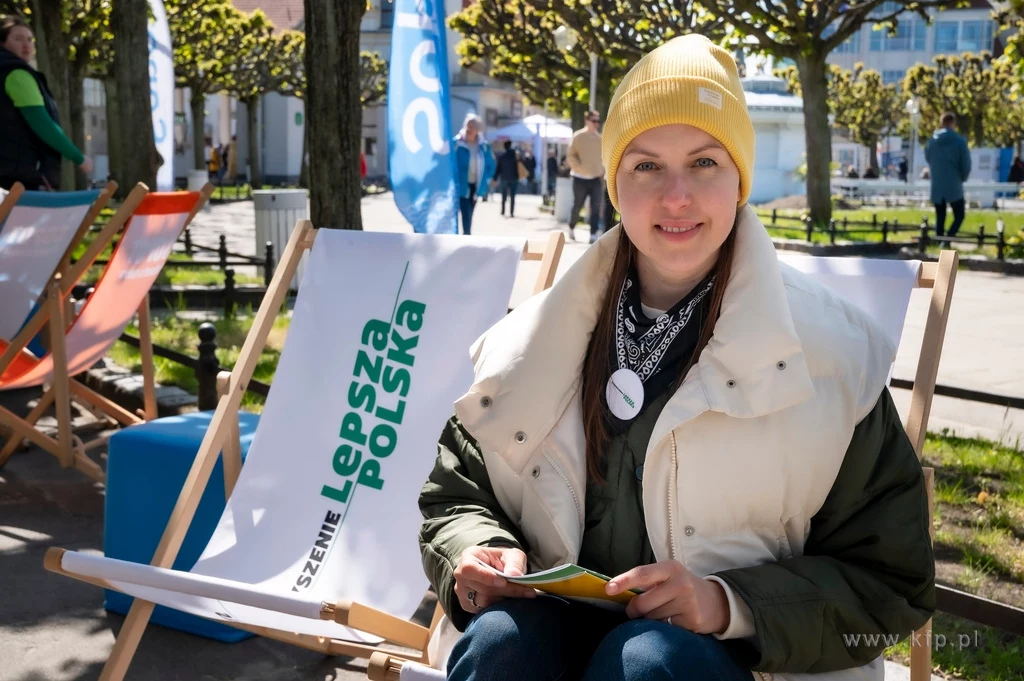 Czyste, Zielone Miasta w Sopocie fot. Anna Bobrowska/KFP/UMS Młoda kobieta w żółtej czapce siedzi na leżaku na zewnątrz, trzymając ulotkę w rękach. Obok niej znajduje się leżak z napisem 'Lepsza Polska'. W tle widoczni są inni ludzie oraz zielone drzewa.