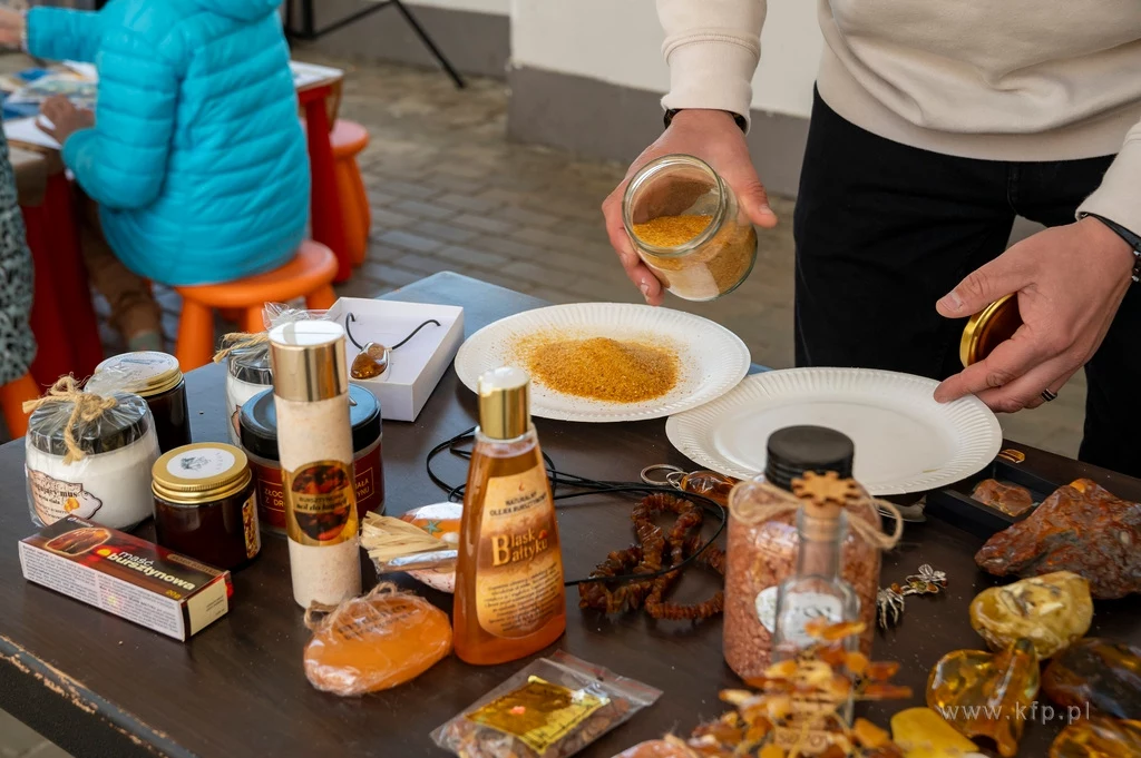 Czyste, Zielone Miasta w Sopocie fot. Anna Bobrowska/KFP/UMS Stół z różnorodnymi produktami, w tym przyprawy, miód i kosmetyki, które są przedstawiane przez osobę w białym swetrze. W tle widać osobę siedzącą na pomarańczowym stołku.