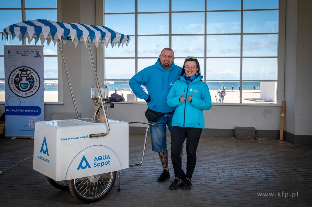Czyste, Zielone Miasta w Sopocie fot. Anna Bobrowska/KFP/UMS Para ubrana w niebieskie bluzy, stoi obok białego wózka z logo Aqua Sopot, w tle widoczny jest plażowy krajobraz z widokiem na morze.