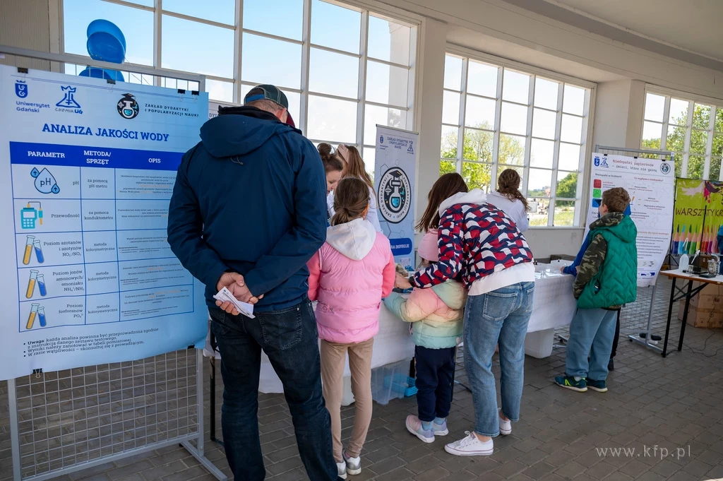 Czyste, Zielone Miasta w Sopocie fot. Anna Bobrowska/KFP/UMS Grupa ludzi oglądająca poster z wynikiem analizy jakości wody. W tle widać dodatkowe tablice informacyjne oraz stoisko edukacyjne za szklanymi oknami. Osoby są ciepło ubrane, co może sugerować chłodniejszą porę roku.