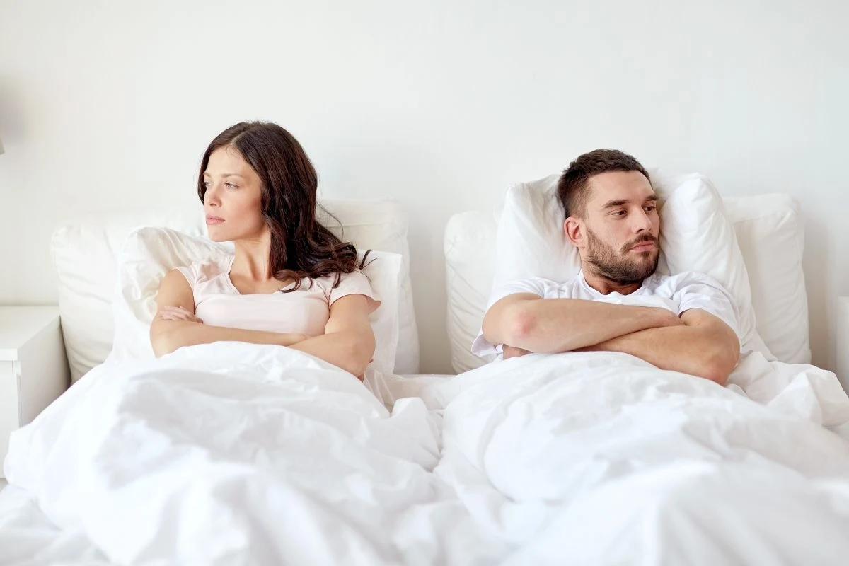 Para siedząca na łóżku w sypialni, oboje odwróceni od siebie z założonymi rękami, co sugeruje napięcie emocjonalne lub konflikt. Jasne wnętrze pokoju tworzy kontrast z ich poważnymi wyrazami twarzy.