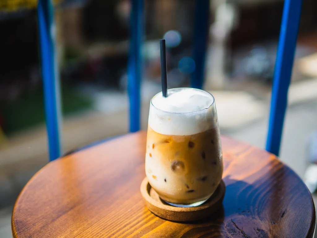 Idealne rozprowadzenie syropu zapewni przystawka do sweet foam Szklanka z mrożoną kawą na drewnianym stoliku w słonecznym otoczeniu. Niebieskie barierki w tle i rozmyte widoki miasta dodają letniego klimatu.