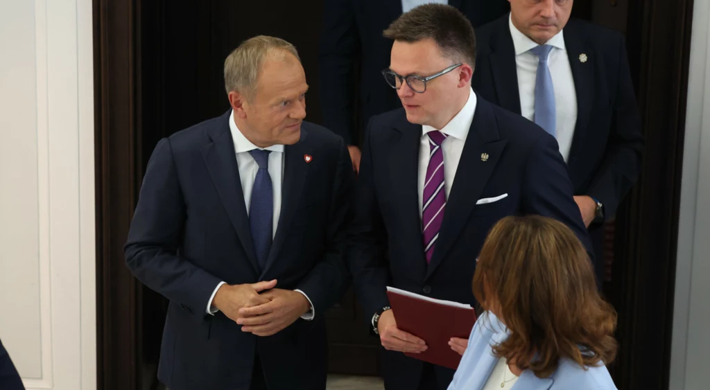 Donald Tusk i Szymon Hołownia Dwóch mężczyzn w garniturach rozmawia w korytarzu, trzymając dokumenty. W tle pojawia się kobieta z długimi włosami oraz jeszcze jeden mężczyzna.