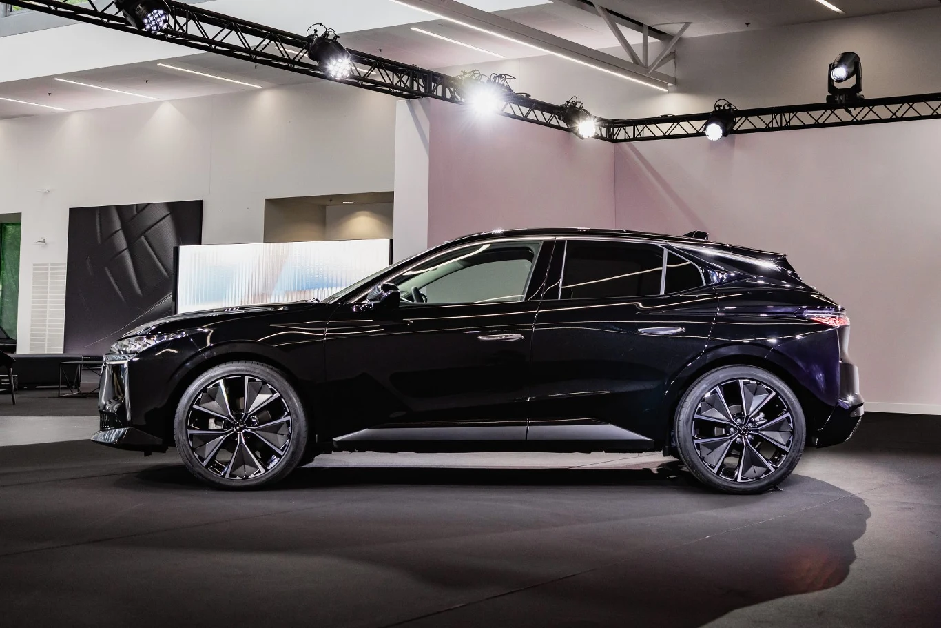 Czarny samochód SUV stoi w nowocześnie oświetlonej sali wystawowej. Pojazd cechuje się nowoczesnym designem i dużymi, stylowymi felgami. W tle widoczne są elementy oświetlenia scenicznego oraz jasne ściany wystawowe.