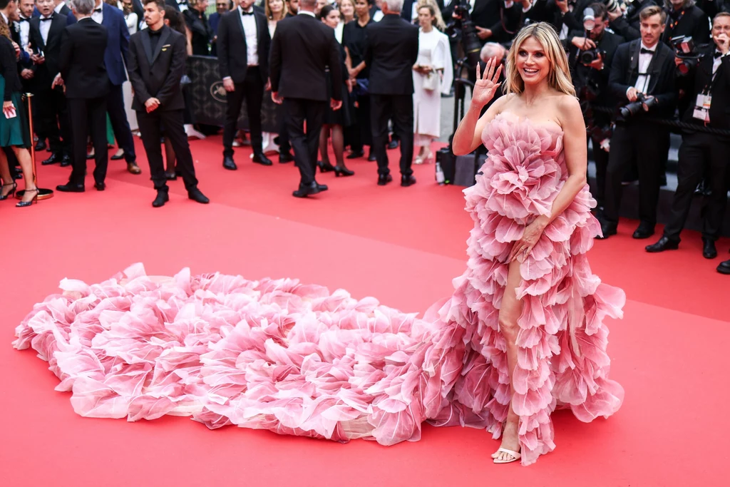 Heidi Klum na 78. Festiwalu Filmowym w Cannes Heidi Klum w różowej sukni na czerwonym dywanie pozdrawia tłum. Suknia jest bogato zdobiona falbanami, które ciągną się za nią, przypominając tren. Obok widać zgromadzonych ludzi ubranych w eleganckie stroje, patrzących w jej kierunku.