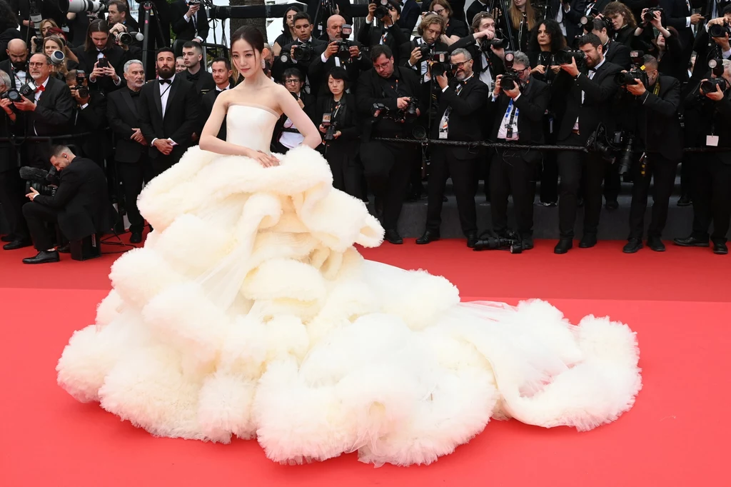 Cannes 2025: Wan Qian Hui Wan Qian Hui w efektownej, białej sukni z długim, falbaniastym trenem na czerwonym dywanie otoczona przez tłum fotografów i widzów.