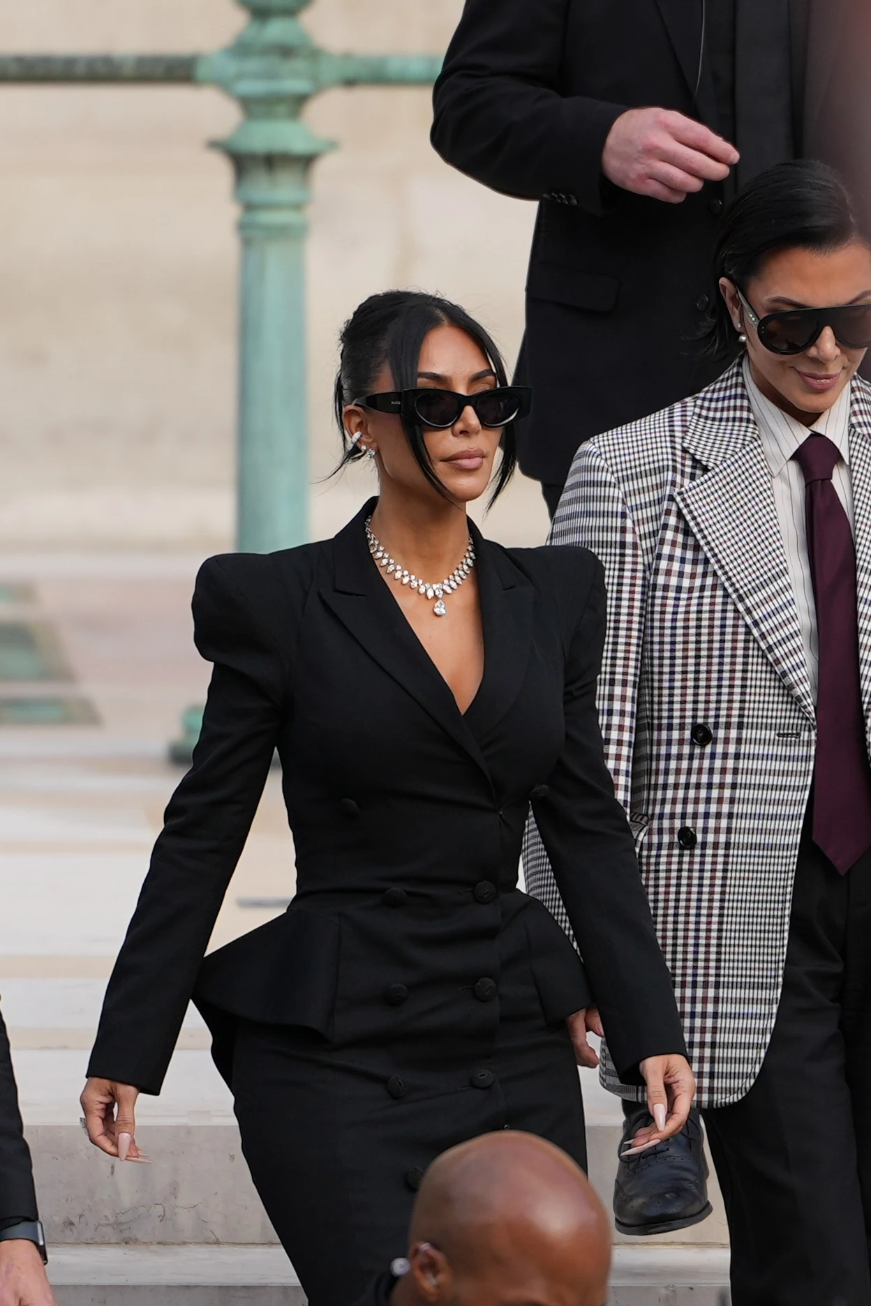 Stylizacja Kim Kardashian i jej biżuteria zwróciły uwagę Mężczyzna i kobieta elegancko ubrani, wychodzący po schodach, on w kraciastej marynarce, ona w czarnym kostiumie z okularami przeciwsłonecznymi i naszyjnikiem.