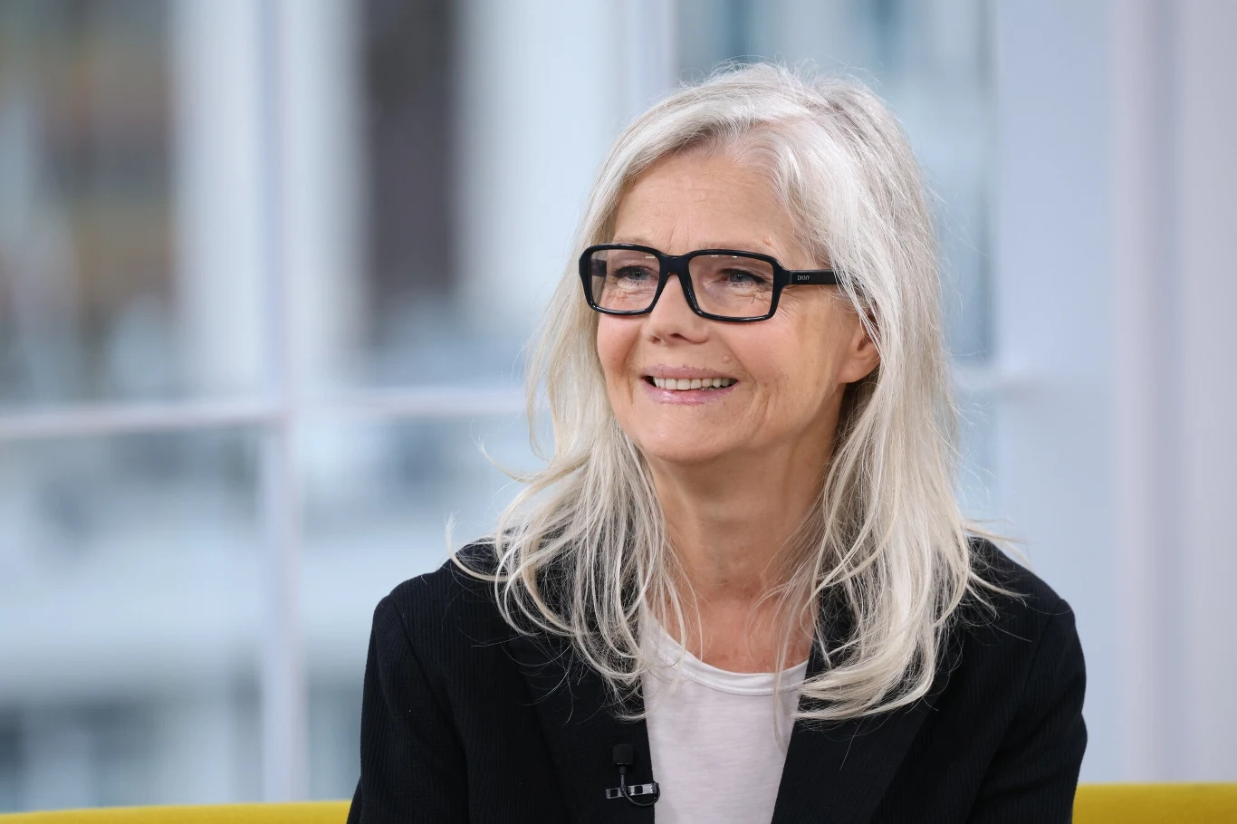 Katarzyna Butowtt była jedną z najbardziej znanych modelek w Polsce w latach 70. i 80. Kobieta w średnim wieku z długimi, siwymi włosami, w okularach i ciemnym marynarce siedzi na żółtej kanapie, uśmiechając się. Za nią widać jasne, rozmyte okno dające dużo naturalnego światła.