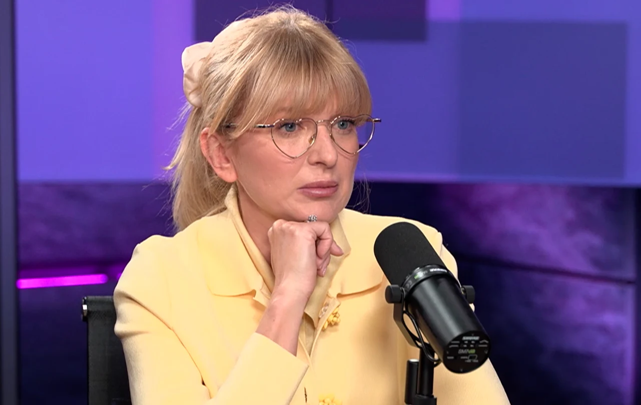 Justyna Szyc-Nagłowska Osoba siedząca w studiu radiowym lub telewizyjnym, ubrana w jasnożółty strój, delikatnie wspiera podbródek na zaciśniętej dłoni. W tle widoczne są elementy sprzętu nagraniowego oraz oświetlenie.