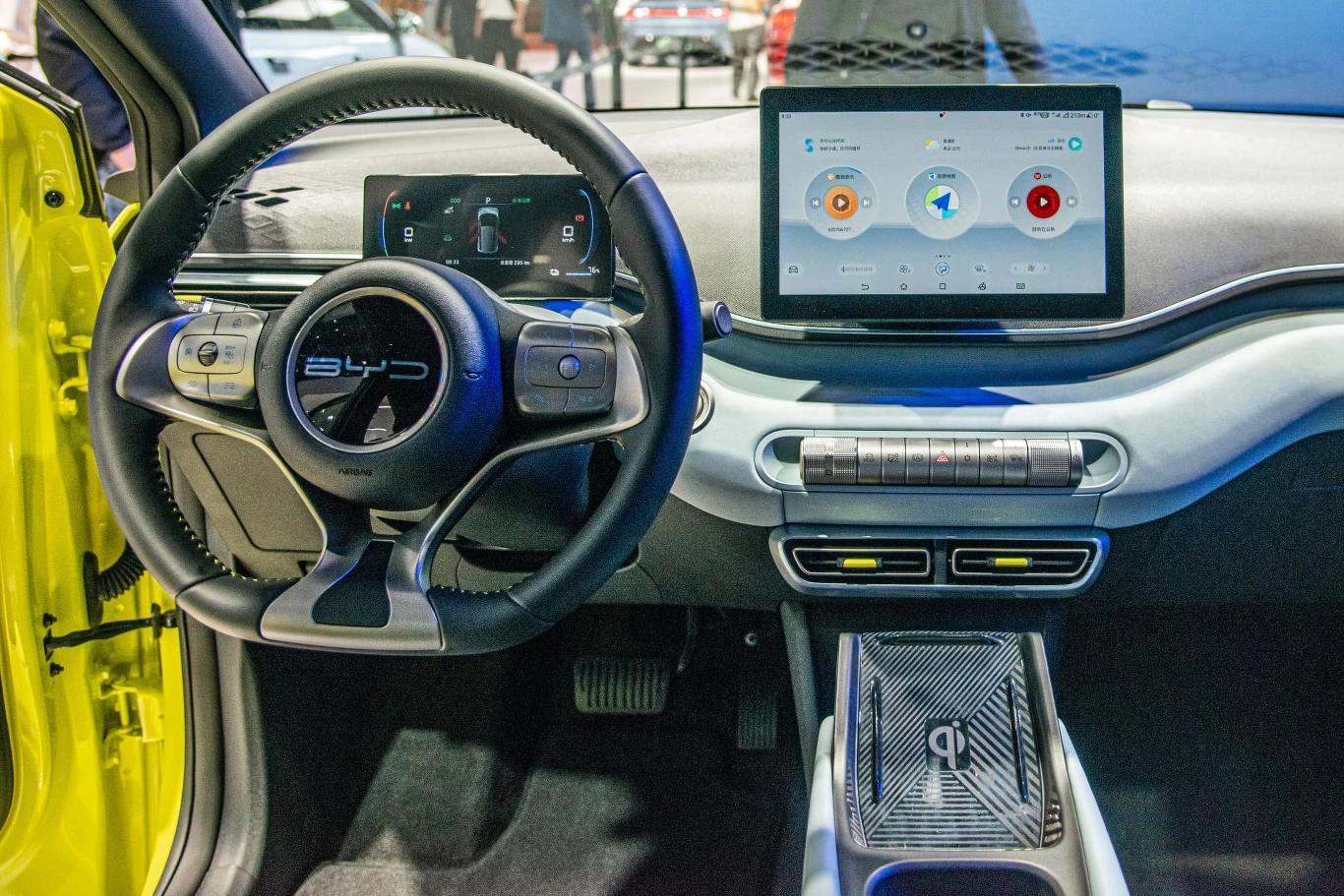 Wnętrze nowoczesnego samochodu elektrycznego, z centralnym dużym ekranem dotykowym oraz kierownicą marki BYD. Ekran wyświetla informacje dotyczące wydajności pojazdu. Stylowy design kokpitu z nowoczesnymi akcentami.