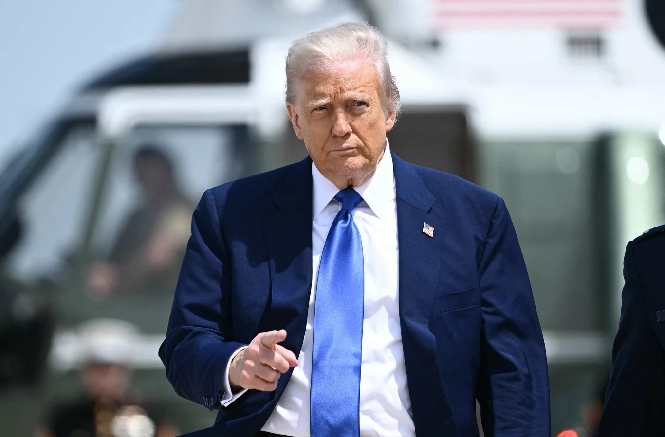 Donald Trump Mężczyzna w granatowym garniturze i niebieskim krawacie, wskazuje palcem w kierunku aparatu. W tle znajduje się helikopter.