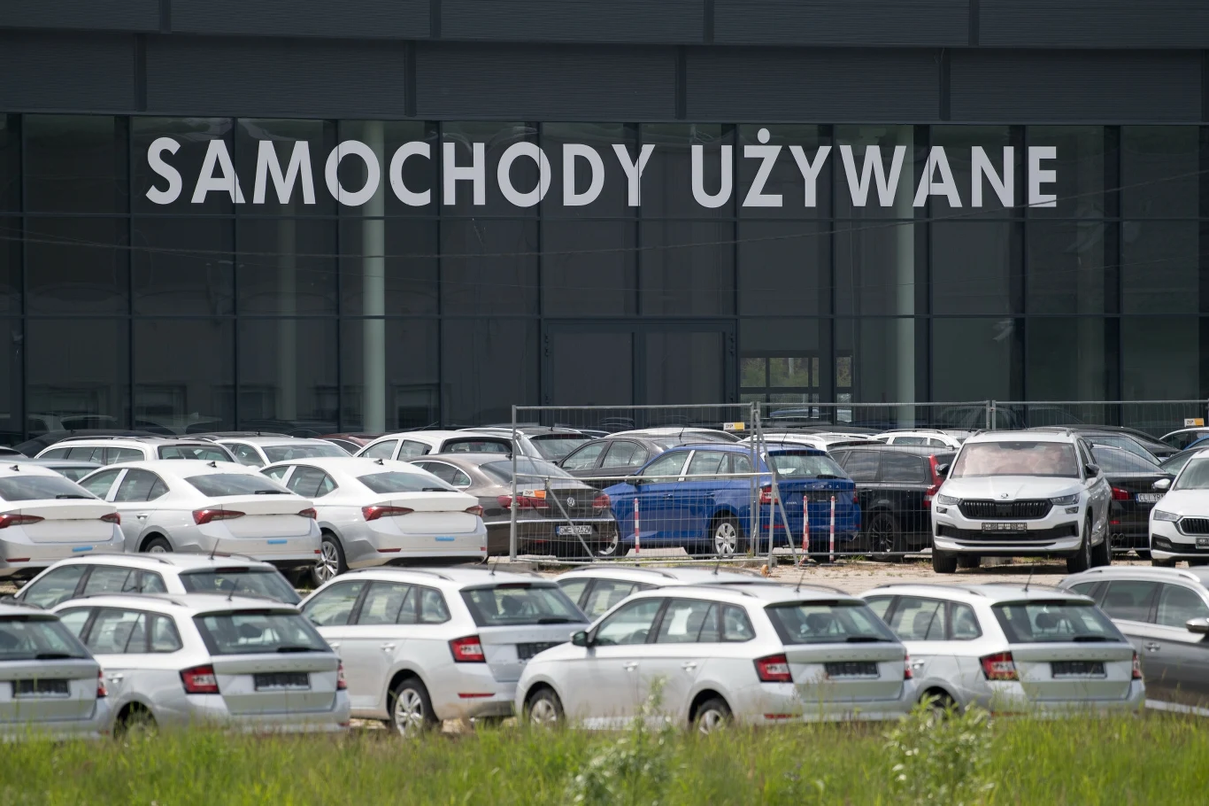 Statystyczny samochód używany w Polsce kosztował w maju 2025 roku już ponad 46 tys. zł. To prawie trzykrotnie więcej niż 10 lat temu Szereg używanych samochodów zaparkowanych przed budynkiem z dużym napisem 'SAMOCHODY UŻYWANE', co sugeruje, że jest to salon sprzedaży samochodów używanych.