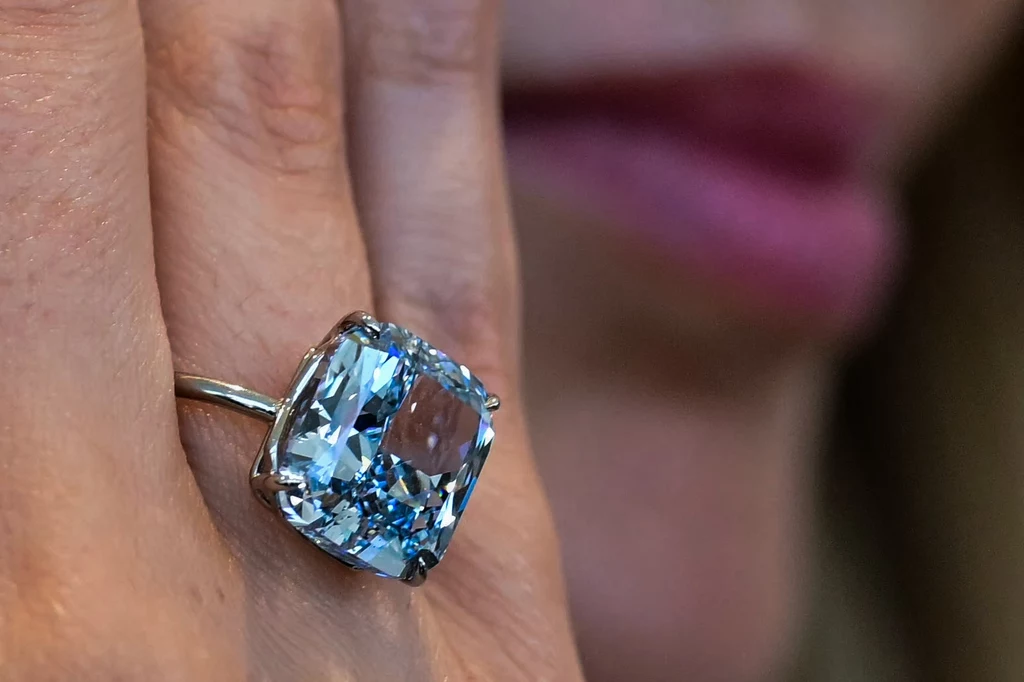 Pierścionek z 10,03-karatowym diamentem "The Mediterranean Blue” cieszył się dużym zainteresowaniem oferentów. Wcześniej był prezentowany na wystawie. lśniący niebieski diamentowy pierścionek na dłoni, w tle niewyraźny zarys twarzy kobiety