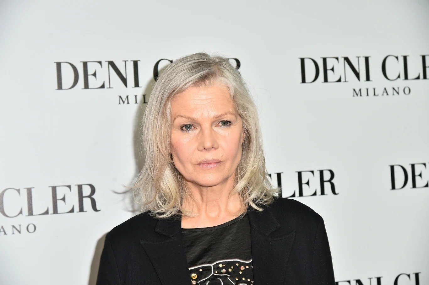 Katarzyna Butowtt od lat udziela się w Fundacji Dr Clown Kobieta z jasnymi włosami na tle ściany z logo marki Deni Cler Milano, ubrana w czarny żakiet i ciemną bluzkę.