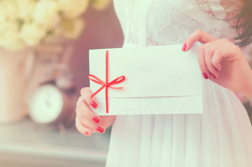 Kobieta w białej sukience trzyma w dłoniach elegancką, białą kopertę przewiązaną czerwoną wstążką. W tle widoczne są rozmyte kwiaty i zegar, co dodaje zdjęciu romantycznego charakteru.