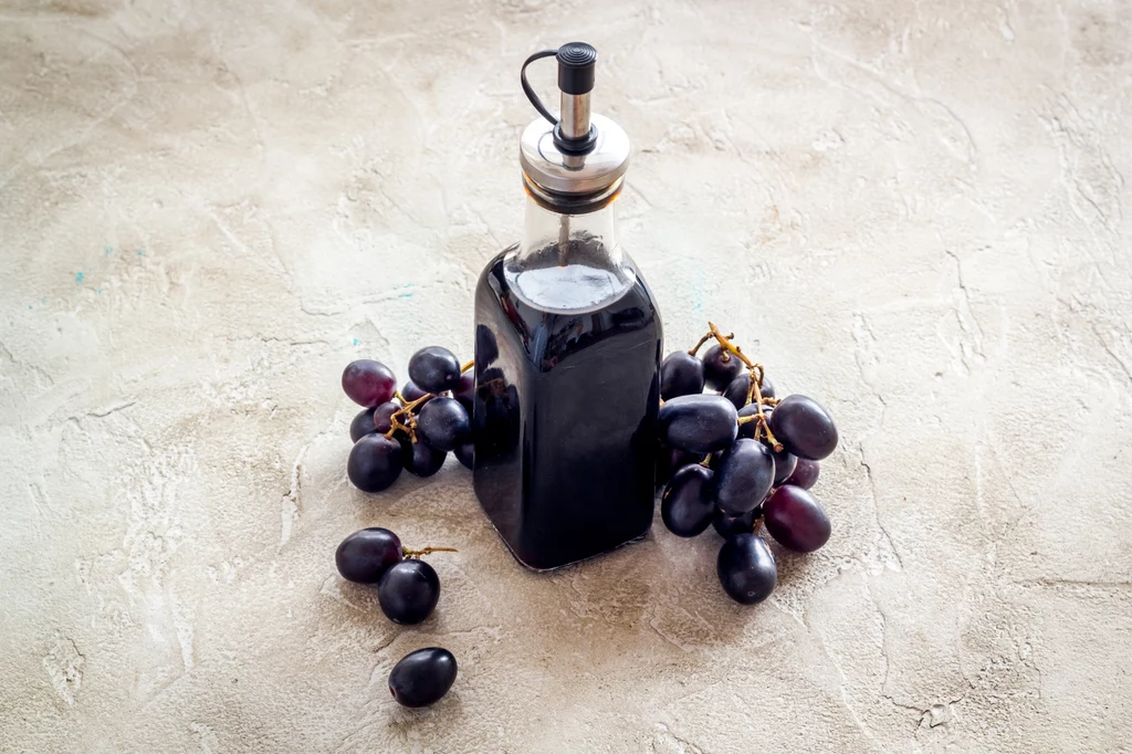 Ocet balsamiczny powstaje z soku winogron Butelka z oliwą z oliwek na jasnym tle otoczona ciemnymi, soczystymi winogronami.