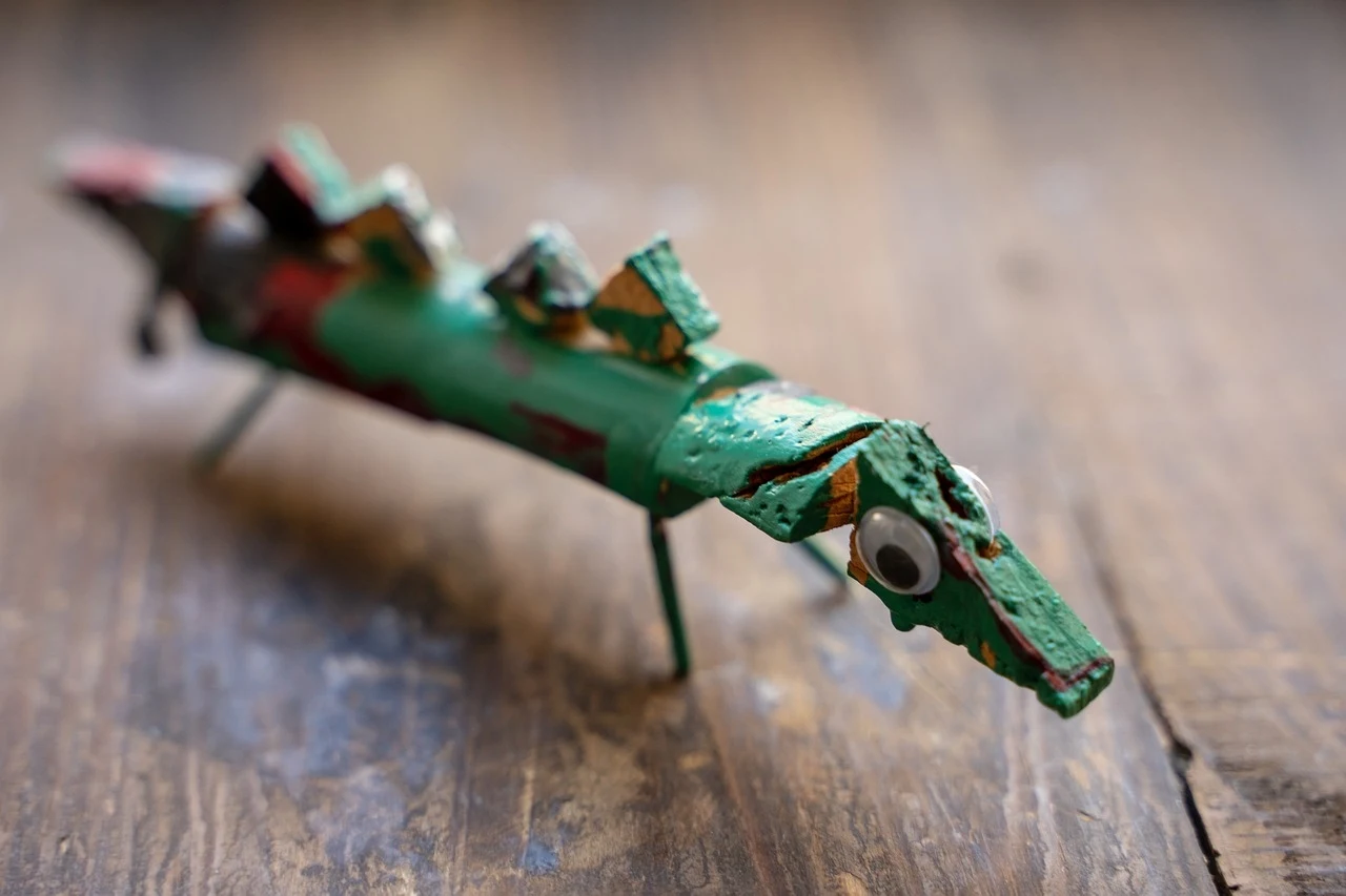 Drewniana figurka o zielonej i czerwonej kolorystyce przypominająca krokodyla, z dużym okiem i nierównymi detalami. Spoczywa na drewnianej powierzchni, co podkreśla jej rustykalny charakter.