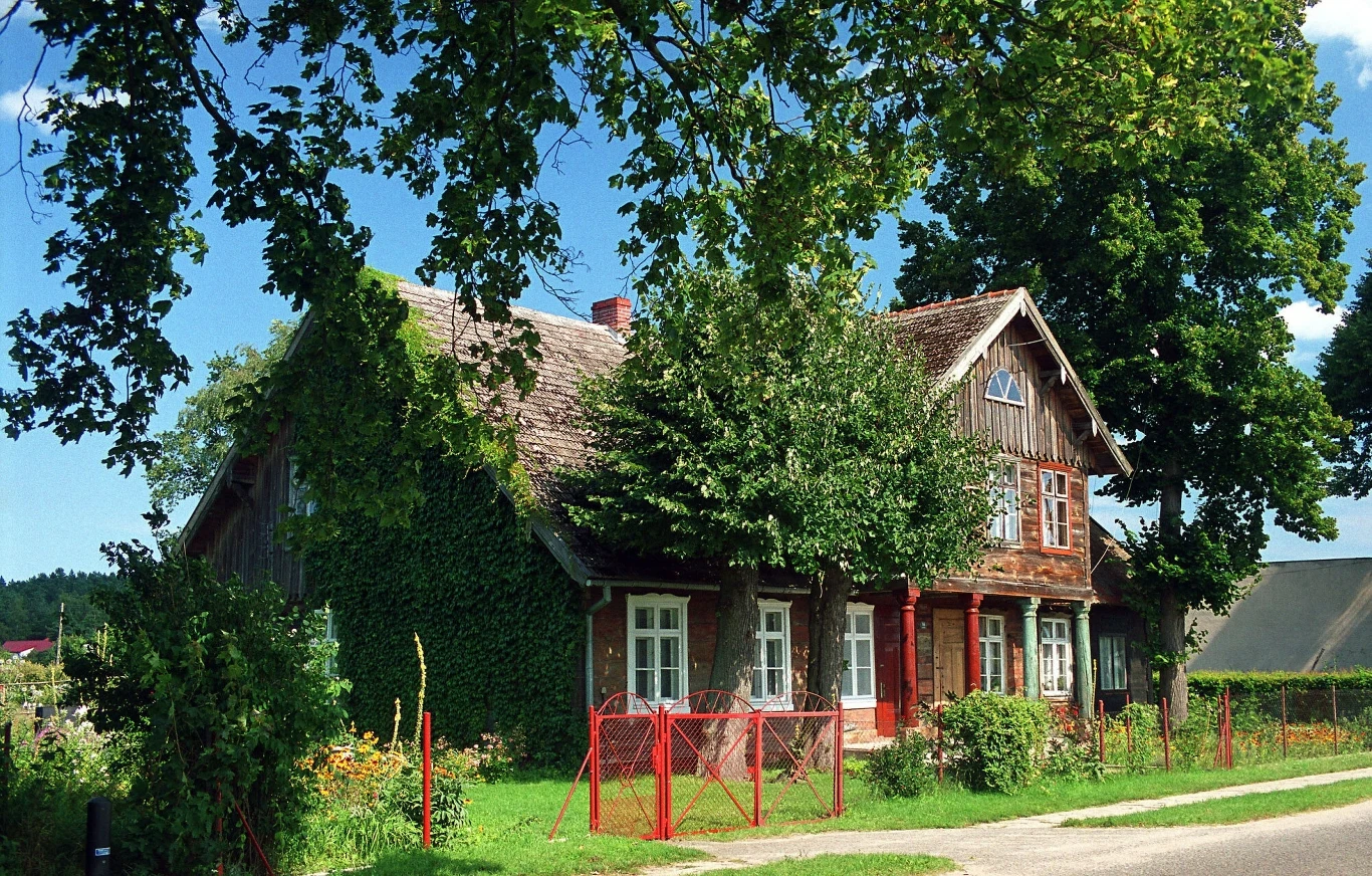 Drewniany, wiejski dom otoczony bujną roślinnością i drzewami. Przed budynkiem znajduje się czerwone ogrodzenie, a całość emanuje spokojem i nostalgią.