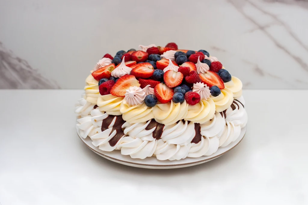Zamiast kremu z mascarpone możesz przygotować budyniowy krem patissiere i polać bezę sosem czekoladowym Tort z białymi bezowymi warstwami udekorowany świeżymi owocami jak truskawki, maliny i jagody oraz bitą śmietaną.