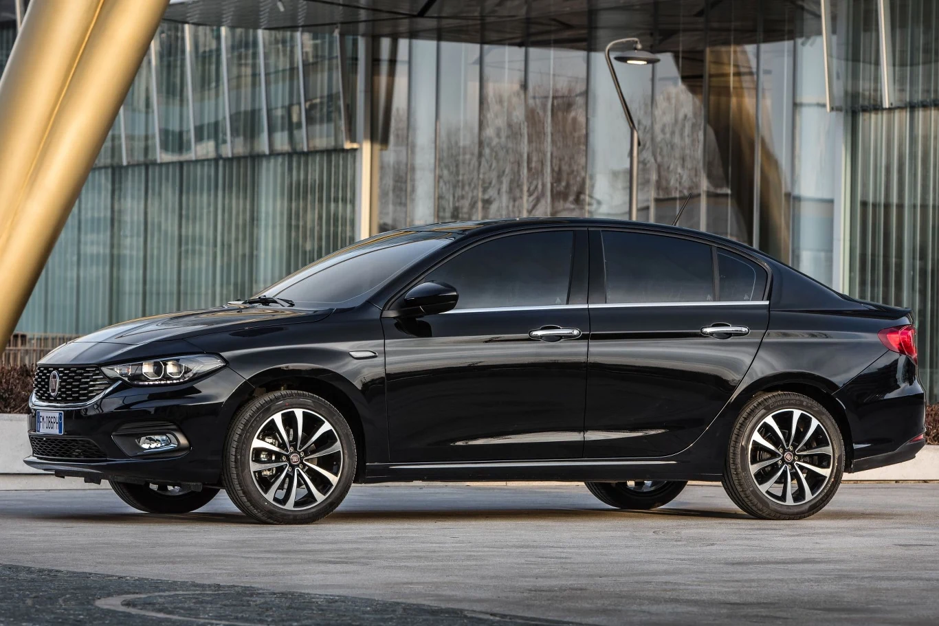 Czarny sedan zaparkowany przed nowoczesnym budynkiem ze szklanymi ścianami i złotymi filarami. Samochód ma ciemne szyby i felgi aluminiowe, stoi na betonowej nawierzchni.