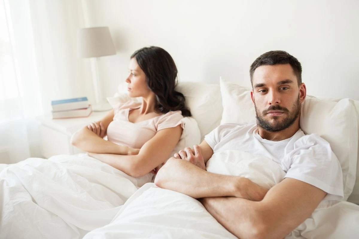 Para siedząca w łóżku, oboje wyglądają na zamyślonych lub nieco zirytowanych. Kobieta patrzy w bok, a mężczyzna patrzy prosto przed siebie. Oboje mają skrzyżowane ręce, co może sugerować napiętą atmosferę.