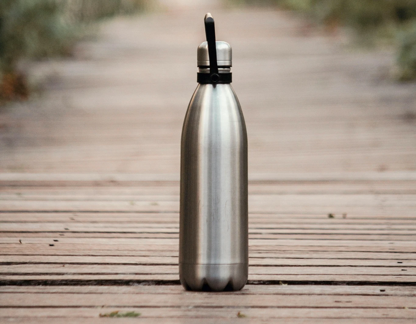 Stalowa butelka z czarnym uchwytem stoi na drewnianej ścieżce. Ścieżka rozciąga się w tle, prowadząc przez zalesiony teren.