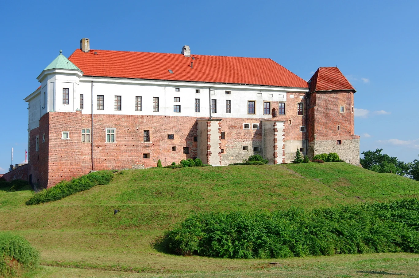 Zamek na wzgórzu o czerwonym dachu otoczony zielenią. Budynek z cegły z białymi akcentami, charakterystyczny dla architektury renesansowej, prezentuje solidną bryłę i masywne mury. Tło wypełnia błękitne niebo.