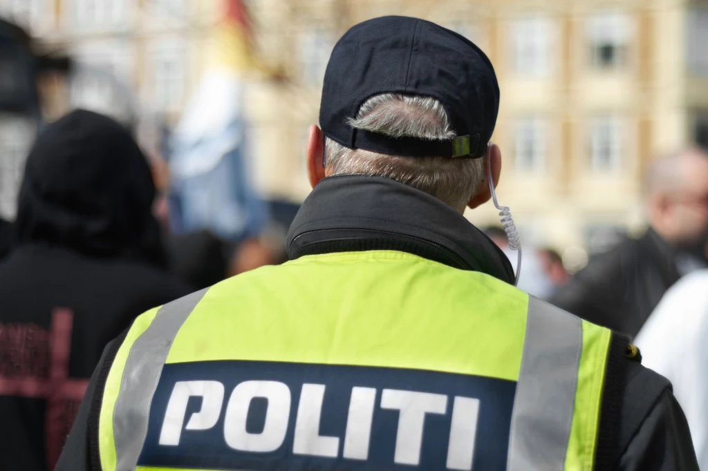 Protesty często zamieniały się w potyczki z policją. Policjant w jaskrawej kamizelce z napisem 'POLITI' patrzący w stronę tłumu. Tło zamazane, co wskazuje na sytuację miejską, prawdopodobnie podczas wydarzenia publicznego lub manifestacji.